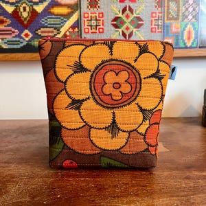 May include: A quilted fabric pouch featuring a large, bold floral pattern in shades of orange, yellow, and brown. The rectangular bag has a red zip closure at the top and a small light blue tag on the side with a black bird silhouette icon.