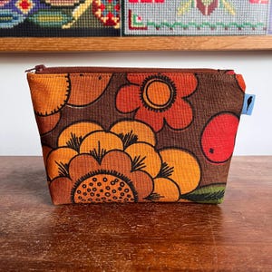 Puede incluir: Un estuche rectangular de tela con cremallera que presenta un fondo marrón con un patrón floral a gran escala en tonos naranja, amarillo y rojo. El estuche tiene un cierre de cremallera marrón oscuro en el borde superior y una pequeña etiqueta azul claro.