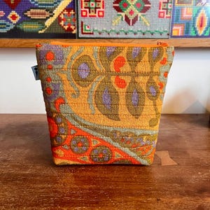 Könnte beinhalten: Ein gestepptes Stofftäschchen mit einem lebendigen Paisleymuster in Orange, Senfgelb, Violett und Salbeigrün. Das Täschchen hat eine horizontal gesteppte Textur, einen orangefarbenen Reißverschluss an der Oberseite und ein kleines graues Etikett.