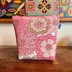 May include: A zipped pouch featuring a woven fabric with a repeating floral pattern in shades of pink, light pink, and beige. The rectangular bag has a pink zip closure at the top and a small blue rectangular tag on the side with a black bird icon.