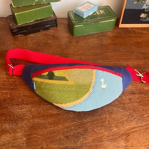 May include: A bum bag featuring a needlepoint front panel with a green, yellow, and light blue geometric design. The bag has a dark blue fabric body, a bright red adjustable strap with a metal buckle, and a red zipper closure across the top.