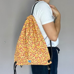 Può includere: Una borsa a cordoncino gialla con un motivo floreale con fiori rossi e gialli. La borsa ha laccetti neri e un piccolo cartellino blu con un logo.