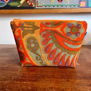 May include: A zipped pouch featuring a vibrant, textured fabric with a bold paisley and floral pattern in shades of bright orange, muted olive green, brown, and soft lavender. The top edge is finished with a matching orange zip closure.