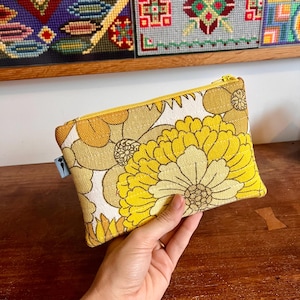 May include: A rectangular zipped pouch made of textured fabric featuring a large-scale floral pattern in shades of yellow, mustard, and white. The pouch has a yellow zip closure and a small light blue side tag with a black carrot icon.