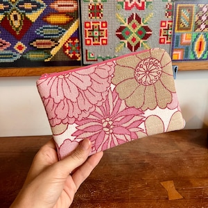 May include: A rectangular zipped pouch featuring a woven fabric pattern of large stylised flowers in shades of pink, light pink, and tan. The pouch has a bright pink zip closure along the top edge and a small blue fabric tab on the side.