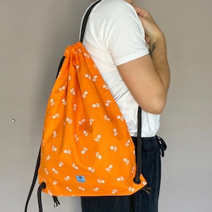 May include: Orange drawstring bag with a white floral pattern. The bag has black drawstrings and a small blue label with a white logo.