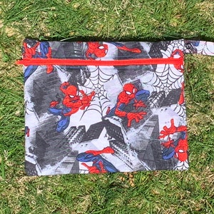 May include: A red and blue Spiderman themed zippered pouch with a red zipper and a black handle. The pouch is made of a white fabric with a black and white comic book style background. The pouch is lying on a green grassy surface.