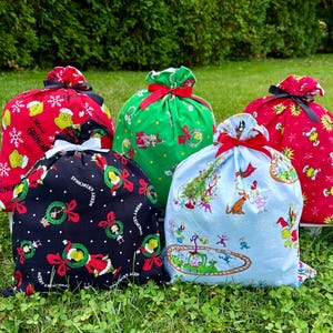 May include: Five Christmas-themed drawstring gift bags in various colors. The bags feature Grinch characters and holiday designs on red, green, black, and light blue fabrics. Each bag is tied with a contrasting ribbon.