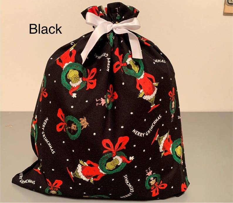 May include: Black fabric gift bag with a white ribbon closure. The bag is patterned with a repeating design of a green Grinch character wearing a red Santa suit and holding a wreath. The words "Merry Grinchmas" are also printed on the bag.