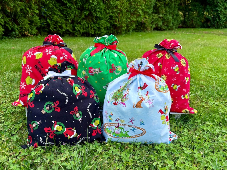 May include: Five Christmas-themed drawstring gift bags in various colors. The bags feature Grinch characters and holiday designs on red, green, black, and light blue fabrics. Each bag is tied with a contrasting ribbon.