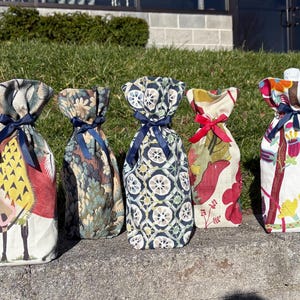 May include: Five fabric wine bottle bags with various patterns and colors. The bags are tied at the top with ribbons. The patterns include floral, geometric, and bird designs. The bags are standing on a concrete surface.