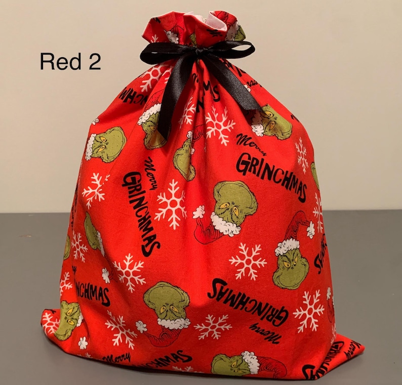 May include: Red Christmas gift bag with a black ribbon closure. The bag is covered in a pattern of the Grinch, snowflakes, and the words "Merry Grinchmas". The text "Red 2" is in the upper left corner.