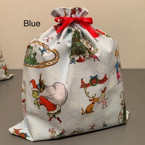 May include: A light blue fabric gift bag with a red ribbon closure. The bag is decorated with colorful illustrations of characters and scenes from a holiday story. The word "Blue" is printed on the bag.