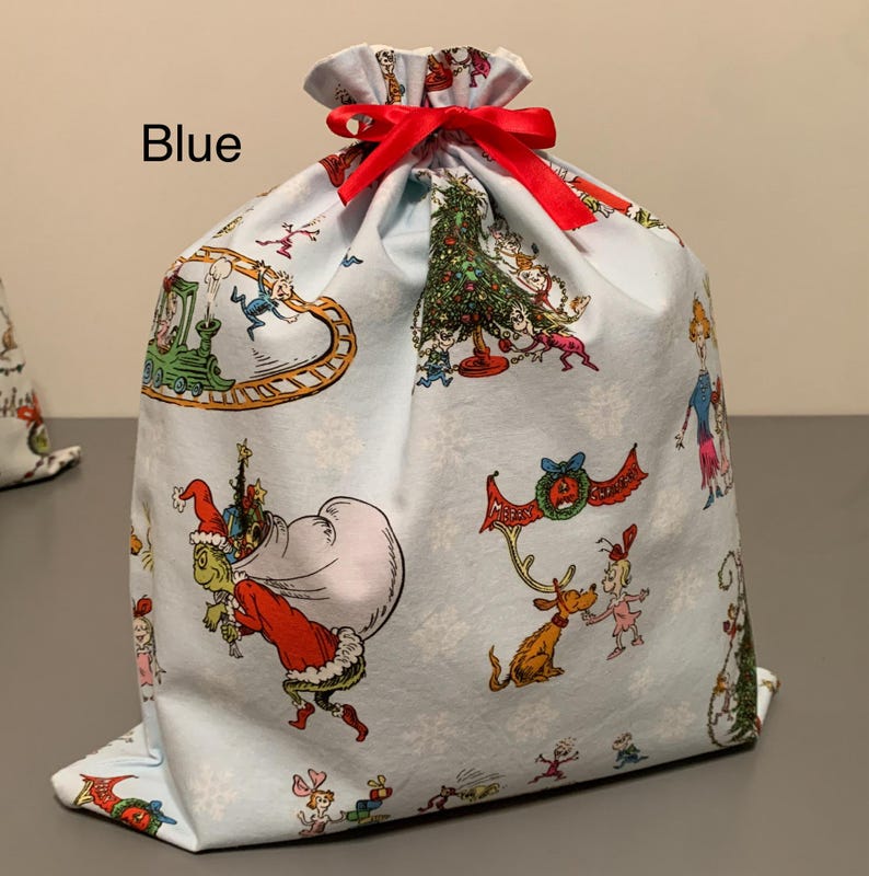 May include: A light blue fabric gift bag with a red ribbon closure. The bag is decorated with colorful illustrations of characters and scenes from a holiday story. The word "Blue" is printed on the bag.