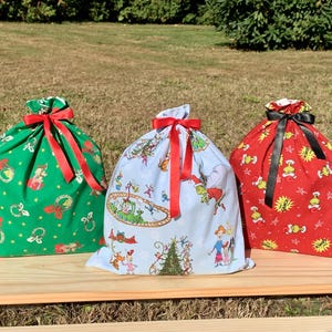 May include: Three Christmas-themed fabric gift bags with drawstring closures and red or black ribbon bows. The bags are green, light blue, and red, each with a different festive pattern. They sit on a wooden surface.
