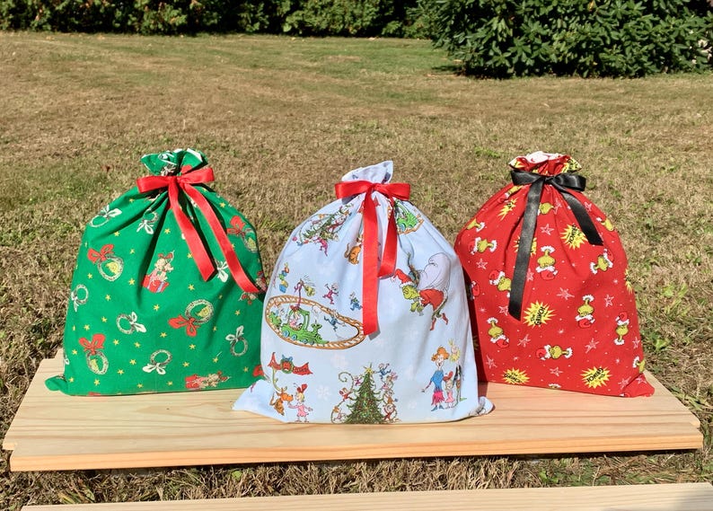 May include: Three Christmas-themed fabric gift bags with drawstring closures and red or black ribbon bows. The bags are green, light blue, and red, each with a different festive pattern. They sit on a wooden surface.