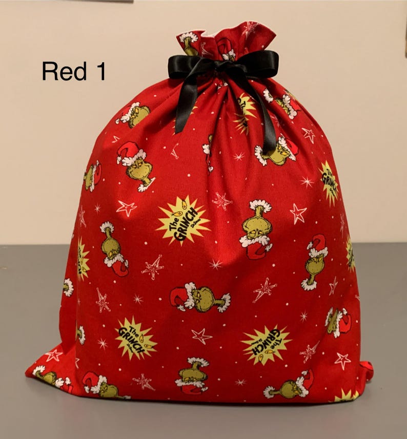 May include: Red drawstring gift bag with a black bow. The bag is covered in a pattern of the Grinch, stars, and text that says "The Grinch". The text "Red 1" is in the upper left corner.