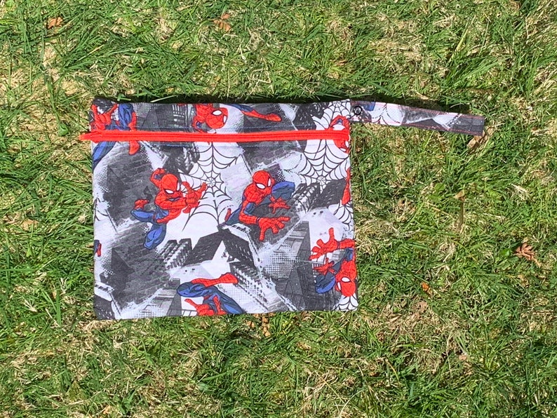 May include: A red and blue Spiderman themed zippered pouch with a red zipper and a black handle. The pouch is made of a white fabric with a black and white comic book style background. The pouch is lying on a green grassy surface.