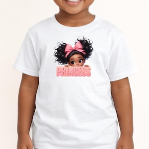 Può includere: Una t-shirt bianca a maniche corte con la grafica di un giovane personaggio dai capelli scuri ricci acconciati in due chignon con un fiocco rosa. Il personaggio spunta sopra la parola PRINCESS scritta in lettere rosa con un bordo chiaro.