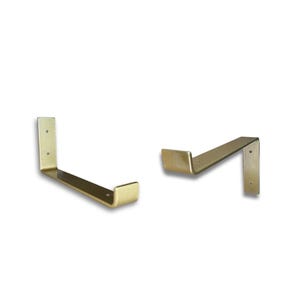 May include: Two gold-coloured metal shelf brackets. Each bracket has a right-angle shape with a flat horizontal support and a vertical mounting plate. The brackets are designed for wall mounting and are suitable for supporting shelves.