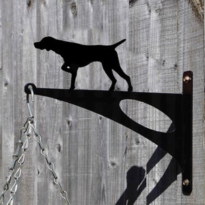 May include: Black metal garden hanger featuring a silhouette of a German Shorthaired Pointer dog. The hanger is mounted on a wooden fence and has a chain hanging from it.