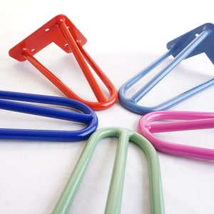 May include: A selection of hairpin table legs in various colours: red, blue, light blue, green, and pink. Each leg features a flat mounting plate with pre-drilled holes and a three-rod design. These metal legs are suitable for DIY furniture projects.