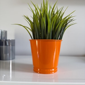Op de afbeelding: Een feloranje plantenpot gevuld met kunstgras. De glanzende, cilindrische pot heeft een licht uitlopende basis en staat op een wit oppervlak. Het kunstgras heeft lange, smalle bladeren.