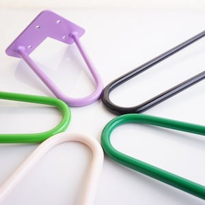 May include: A selection of hairpin table legs in a range of colours: green, purple, black, and off-white. The metal legs have a curved, U-shape design. The purple leg features a mounting plate with pre-drilled holes for easy attachment.