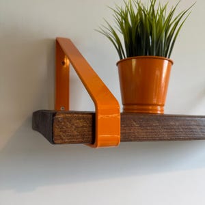Op de afbeelding: Een zwevend plankje met een donker gebeitste houten plank en een feloranje metalen beugel. Een oranje metalen pot met groen gras staat op de plank. De plank is aan een witte muur bevestigd.