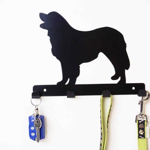 Könnte beinhalten: Schwarzer Schlüsselhaken aus Metall in Form einer Hundesilhouette. Der Haken hat drei Haken zum Aufhängen von Schlüsseln oder Leinen. Ein blauer Schlüsselanhänger und eine grüne Leine hängen an den Haken.