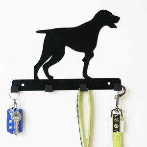 May include: Black metal key hook shaped like a dog silhouette. The hook has three hooks for hanging keys or leashes. A green leash and a silver key chain with keys are hanging on the hooks.