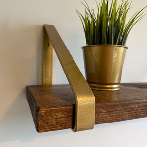 Op de afbeelding: Een donkerbruine houten zwevende plank ondersteund door een goudkleurige metalen beugel. Een kleine goudkleurige metalen pot met een kunstmatige grasplant staat op het oppervlak van de plank. De plank is gemonteerd tegen een effen witte muur.