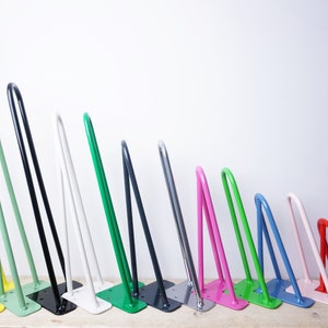May include: A row of 14 hairpin table legs in various colours, including yellow, green, black, white, pink, blue, and red. The legs are made of metal and have a simple, modern design.