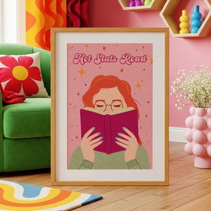 May include: A framed wall art print featuring a stylized illustration of a person with red hair and glasses reading a dark pink book. The background is light pink with yellow stars and the text "Hot Sluts Read" in a retro, cursive font.