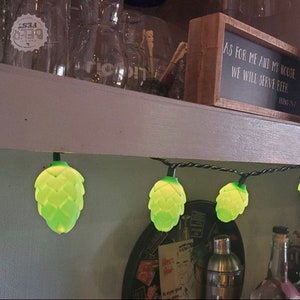May include: A string of decorative lights featuring green hop-shaped bulbs. The lights are strung along a black wire and are illuminated. A sign above the lights reads "AS FOR ME AND MY HOUSE, WE WILL SERVE BEER".