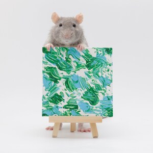 Pu&ograve; includere: Un ratto grigio si trova su un piccolo cavalletto di legno tenendo una tela dipinta con vortici astratti verdi e blu.