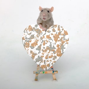 Pode incluir: Um pequeno rato cinzento está atrás de uma tela em forma de coração com um padrão abstrato de salpicos de tinta em dourado metálico, prateado e branco. A tela em forma de coração repousa sobre um cavalete de madeira com manchas de tinta.