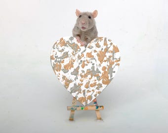 May include: A small grey rat stands behind a heart-shaped canvas featuring an abstract paint splatter pattern in metallic gold, silver, and white. The heart canvas rests on a miniature wooden easel with visible paint stains on its frame.