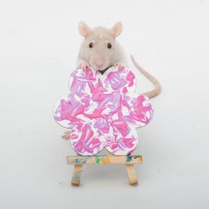Pode incluir: Um rato de cor clara está atrás de um pequeno cavalete de madeira segurando um recorte em forma de flor. O recorte apresenta um design de pintura abstrata em tons de rosa brilhante, rosa claro e branco. O cavalete tem respingos de tinta.