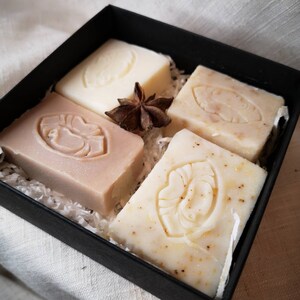May include: Four bars of handmade soap in a black box. The soaps are white and beige with different designs. The box is filled with shredded paper.