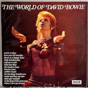 May include: A vintage vinyl record album cover featuring David Bowie in a red and black outfit. The album title "The World of David Bowie" is printed in white letters at the top of the cover. The album cover also includes a list of song titles printed in white text.