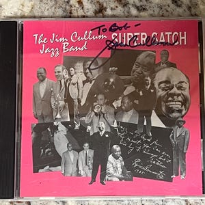 May include: A compact disc case featuring a bright pink cover with black and white photographs of The Jim Cullum Jazz Band. The text reads The Jim Cullum Jazz Band SUPER CATCH. There is a handwritten signature in black ink across the top section.