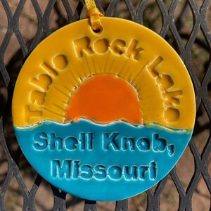 May include: A yellow ceramic ornament with the text "Table Rock Lake Shell Knob, Missouri" and a stylized orange sun with yellow rays.