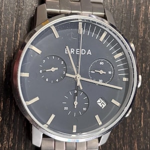 May include: A silver BREDA watch with a dark grey face and a silver strap. The watch face has three sub-dials and a date display. The watch is on a dark wood surface.
