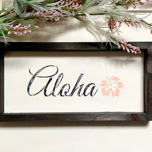 May include: A framed wooden sign with the word "Aloha" in black script and a pink hibiscus flower.