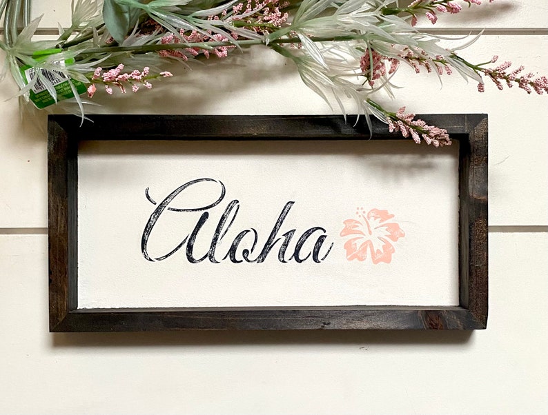 May include: A framed wooden sign with the word "Aloha" in black script and a pink hibiscus flower.