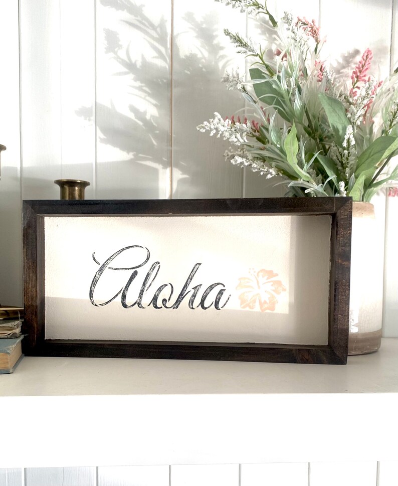 May include: A framed wooden sign with the word "Aloha" in black script and a pink hibiscus flower graphic.