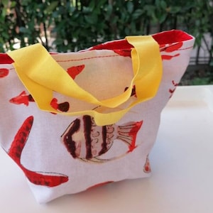Peut inclure: Un petit sac fourre-tout blanc avec des poissons rouges et jaunes imprimés dessus. Le sac a des anses jaunes.