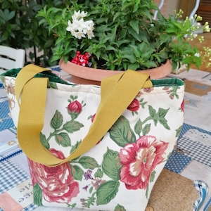 Könnte beinhalten: Eine kleine Blumentasche mit weißem Hintergrund und rotem, rosa und grünem Blumendruck. Die Tasche hat zwei gelbe Henkel.