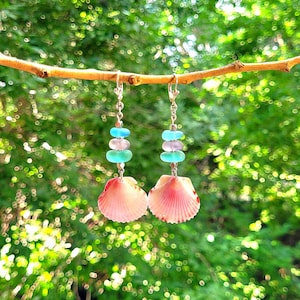 Könnte beinhalten: Ein Paar Hängeohrringe mit rosa Muscheln und blauen und grünen Glasperlen. Die Ohrringe hängen an einem Zweig.